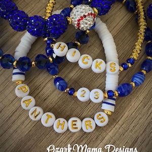 May include: Several beaded bracelets in shades of blue and gold. One bracelet features white letter beads spelling "PANTHERS" and "HICKS 23". A baseball-themed bead is also visible. The bracelets are arranged on a wooden surface. The text "Ozark Mama Designs" is at the bottom.