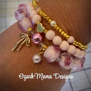 Puede incluir: Una pila de pulseras en tonos rosa y dorado. Las pulseras presentan varias cuentas, incluyendo perlas, cuentas rosas mate y cuentas de vidrio decorativas con patrones florales. Los dijes dorados incluyen un lazo y un corazón. El texto "Ozark Mama Designs" es visible.