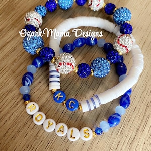 May include: Three beaded bracelets in shades of blue, white, and gold. One bracelet spells out "ROYALS" in gold letters. Other bracelets feature baseball-themed beads and sparkling blue beads. The text "Ozark Mama Designs" is visible.