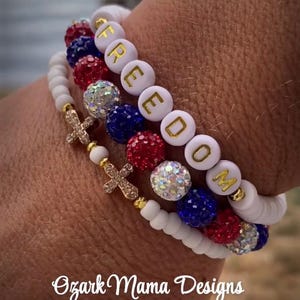 Könnte beinhalten: Drei Armbänder mit roten, weißen und blauen Perlen. Ein Armband schreibt das Wort "FREEDOM" in goldenen Buchstaben. Ein weiteres Armband hat goldene Kreuzanhänger. Die Armbänder haben ein patriotisches Thema.