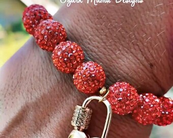 May include: A red beaded bracelet with a gold heart-shaped padlock clasp. The beads are round and covered in sparkling red crystals. The text "Ozark Mama Designs" is visible in the background.