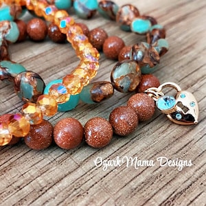 May include: Close-up of three beaded bracelets. One bracelet features orange crystal beads, another has brown and turquoise beads, and the third has brown beads and a gold heart-shaped charm with a blue gem. The bracelets are on a wooden surface.