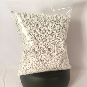 May include: A clear plastic bag filled with white perlite, a horticultural growing medium. The bag is sitting on a black, rounded container. The perlite appears to be small, irregular pieces, and the bag is sealed at the top.