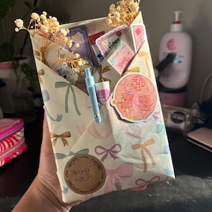 May include: A cream-colored paper envelope decorated with pastel bows, containing dried baby's breath flowers, a light blue pen, and various stickers including a holographic disco ball design. A circular brown sticker reads Young Vault.