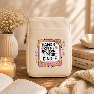 May include: A beige canvas e-reader sleeve with a zipper closure. The front features a graphic of a pink tablet with yellow caution tape and the text: HANDS OFF MY EMOTIONAL SUPPORT KINDLE, accented with small pink hearts and white flowers.