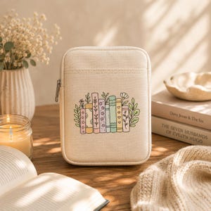May include: A rectangular beige canvas zipped pouch featuring a central graphic illustration of a row of colourful books decorated with floral and heart patterns, surrounded by green leafy sprigs and small flowers on the front exterior.