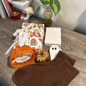May include: A pair of brown fuzzy socks, an orange pumpkin-shaped glass candle labelled Pumpkin Vanilla Brulee, a small gold pumpkin, a white ghost-shaped ceramic dish, a spiral-bound notebook, and a gift wrapped in patterned paper with white lace.