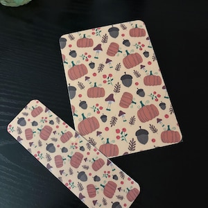 Puede incluir: Una tarjeta rectangular y un marcapáginas largo y estrecho con un patrón repetitivo sobre un fondo crema claro. El diseño incluye calabazas naranjas, bellotas gris oscuro, pequeñas setas moradas y ramitas de bayas rojas.
