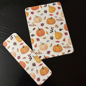 Puede incluir: Una tarjeta rectangular y un marcapáginas largo y estrecho, ambos con fondo blanco y un patrón repetitivo de calabazas naranjas y color crema, varias hojas de otoño en tonos rojos, marrones y amarillos, y pequeñas bayas oscuras.
