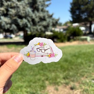 May include: A die-cut sticker featuring an open book with a pair of dark-rimmed glasses resting on the pages, a coffee cup to the left, and pink floral accents surrounding the book, all set against a cloud-shaped background with glitter details.