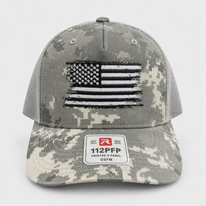 May include: A gray and white digital camouflage trucker hat featuring a distressed, embroidered American flag on the front panel. The cap includes a mesh back and a sticker on the brim that reads: EST. 1970 R 112PFP PRINTED 5 PANEL OSFM.