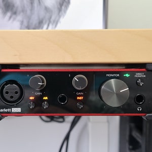 Peut inclure: Une interface audio Scarlett Solo rouge et noire avec deux boutons, un bouton de moniteur et un bouton de moniteur direct. L'interface est montée sous un bureau en bois.