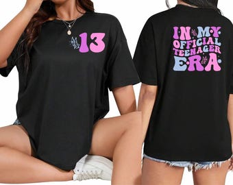May include: Black t-shirt with a pink butterfly and the number 13 on the front. The back of the shirt has the text "In My Official Teenager Era" in pink, blue, and purple letters with butterflies.