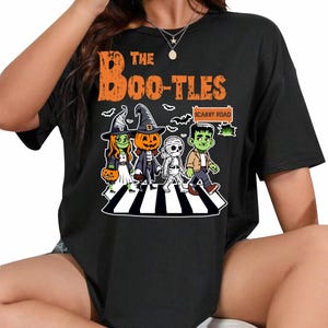 Op de afbeelding: Zwart T-shirt met een afbeelding van een groep Halloween-personages die over een zebrapad lopen. De personages zijn een heks, een pompoen, een mummie en een Frankenstein. De tekst "The Boo-Tles" en "Scabby Road" staan ook op het shirt.
