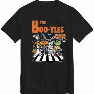 Op de afbeelding: Zwart T-shirt met een afbeelding van vier Halloween-personages die over een zebrapad lopen. De personages zijn een heks, een pompoen, een mummie en een Frankenstein. De tekst "The Boo-Tles" en "Scarby Road" staat op het shirt.