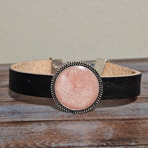 May include: A black leather bracelet with a silver round charm featuring a pink glitter design.