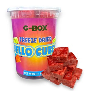 May include: A clear plastic container filled with red freeze-dried jello cubes. The container has a pink label with the text "G-BOX" and "FREEZE DRIED JELLO CUBES". There are also red freeze-dried jello cubes scattered on a white surface.