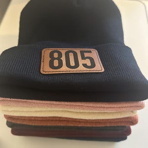 Pu&ograve; includere: Un mucchio di berretti di lana neri, rosa, beige, marroni e bordeaux. Il berretto in cima ha una toppa di pelle marrone con il numero "805" stampato in nero.