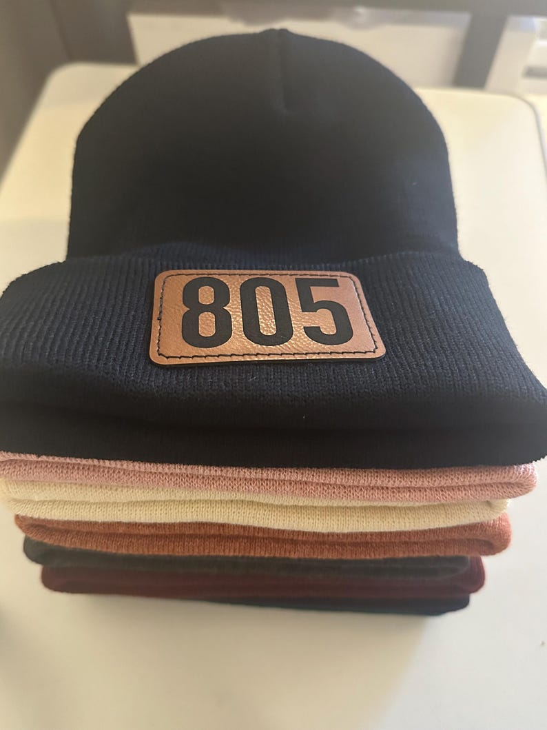 Pu&ograve; includere: Un mucchio di berretti di lana neri, rosa, beige, marroni e bordeaux. Il berretto in cima ha una toppa di pelle marrone con il numero "805" stampato in nero.
