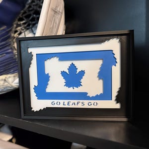 Peut inclure: Une œuvre d'art encadrée représentant le drapeau canadien bleu et blanc avec une feuille d'érable. Les mots "GO LEAFS GO" sont imprimés en bas. L'œuvre est sur une étagère noire, avec un gant de hockey en arrière-plan.