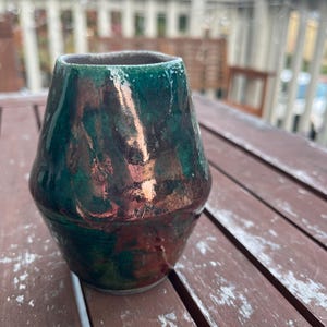Może przedstawiać: Ręcznie wykonany ceramiczny wazon o unikalnym kształcie i błyszczącym wykończeniu. Wazon charakteryzuje się mieszanką zielonych, brązowych i miedzianych kolorów, tworząc efekt marmuru. Wazon stoi na drewnianej powierzchni.