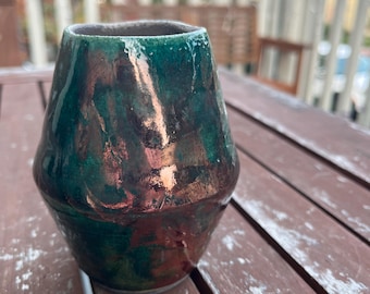 May include: A handmade ceramic vase with a unique shape and a glossy finish. The vase features a blend of green, brown, and copper colors, creating a marbled effect. The vase is sitting on a wooden surface.