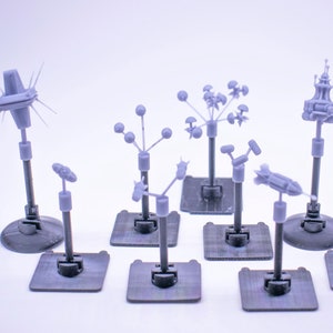 Peut inclure: Une collection de petits modèles gris d'aspect futuriste sur socles. Les modèles présentent divers designs, notamment des sphères, des formes pointues et des structures ressemblant à des fusées ou des antennes. Chaque modèle est monté sur une base gris foncé.