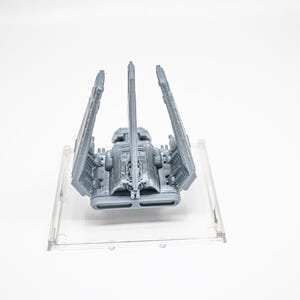 K&ouml;nnte beinhalten: Ein graues 3D-gedrucktes Modell eines Raumschiffs mit drei vertikalen Fl&uuml;geln und einem zentralen Rumpf, montiert auf einer quadratischen, transparenten Kunststoffbasis. Das Schiff zeigt detaillierte Paneele, ein Cockpit und zwei rechteckige Triebwerksausl&auml;sse.