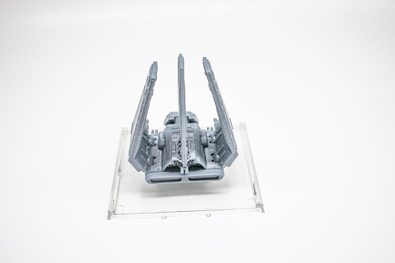 K&ouml;nnte beinhalten: Ein graues 3D-gedrucktes Modell eines Raumschiffs mit drei vertikalen Fl&uuml;geln und einem zentralen Rumpf, montiert auf einer quadratischen, transparenten Kunststoffbasis. Das Schiff zeigt detaillierte Paneele, ein Cockpit und zwei rechteckige Triebwerksausl&auml;sse.