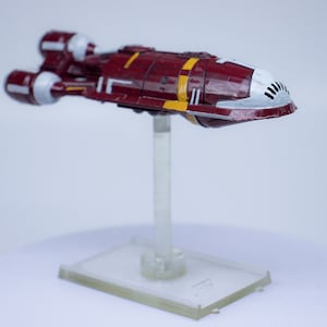 Può includere: Un modello di nave spaziale di colore bordeaux e bianco con accenti gialli su un supporto in plastica trasparente. La nave spaziale ha un lungo naso appuntito e due motori sul retro.