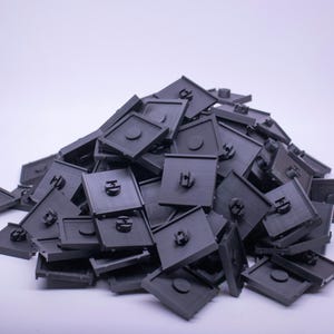 Peut inclure: Un tas de composants carrés en plastique gris foncé, avec un élément circulaire en relief et une petite languette rectangulaire. Les composants sont empilés les uns sur les autres sur fond blanc.