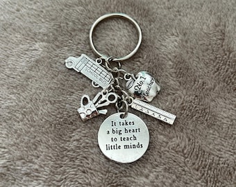 May include: Silver keychain with charms including a school bus, apple, scissors, and a ruler. The charm with the text "It takes a big heart to teach little minds" is the focal point.
