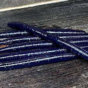 May include: A pile of dark blue fishing worms with a textured surface and subtle glitter accents. The worms are arranged on a weathered wooden surface, suggesting an outdoor or fishing theme.