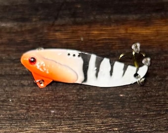 Puede incluir: Un señuelo de pesca con un cuerpo naranja y blanco, rayas negras y un ojo rojo. El señuelo tiene un pequeño labio naranja y un anzuelo triple en la parte trasera. El señuelo está diseñado para atraer peces.