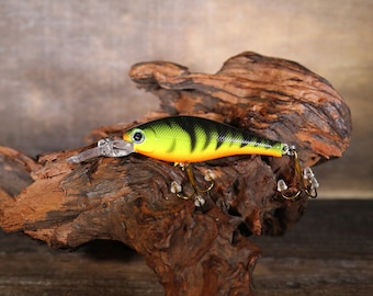 May include: A yellow, black, and orange fishing lure with a realistic fish design. The lure has three treble hooks and is sitting on a piece of driftwood.