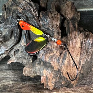 May include: A fishing lure with a red and yellow painted head, a silver spinner blade with black and yellow accents, and a black hook. The lure is attached to a silver wire and has an orange bead.