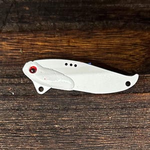 May include: A white, stylized fishing lure with a red eye and a curved body. The lure has a small, white appendage and three small holes. The lure is set against a dark, wooden background.