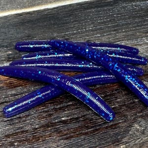 May include: A pile of dark blue, soft plastic fishing lures with blue glitter flecks. The lures are arranged on a weathered, dark wood surface. The lures are cylindrical and slightly curved.