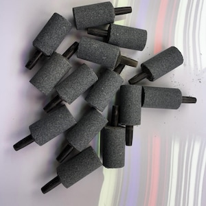 May include: A collection of cylindrical, gray air stones with black stems. These aquarium accessories are designed to diffuse air into water, creating bubbles for oxygenation and aesthetic appeal. The air stones are arranged on a white and multicolored surface.