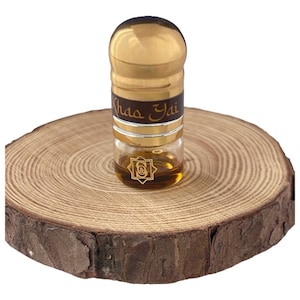 Puede incluir: Un pequeño frasco de perfume de vidrio con tapa dorada y etiqueta marrón con letras doradas, sobre una rodaja de madera. El frasco contiene un líquido marrón dorado. La etiqueta dice "Khada Yai".