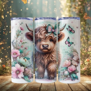 May include: A cylindrical tumbler featuring a brown Highland cow with a teal and pink bow between its horns. The design includes watercolor-style pink and teal flowers, green foliage, and several butterflies against a white background.