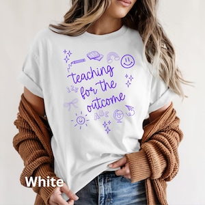 May include: White t-shirt with the text "teaching for the outcome" in purple, along with various doodles like a rainbow, smiley face, and globe. The shirt is a casual style.