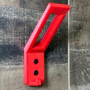 Puede incluir: Soporte de pared rojo brillante, en ángulo, con una abertura rectangular. Dos tornillos negros aseguran el soporte. Diseñado para sujetar un objeto, como una herramienta o equipo.