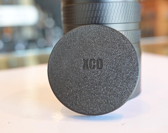 Könnte beinhalten: Eine schwarze Objektivkappe mit dem aufgedruckten Wort "XCO".