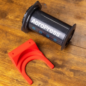 Op de afbeelding: Zwarte AeroPress koffiezetapparaat met witte tekst en blauwe accenten, en een felrode houder. De koffiezetter is cilindrisch en heeft een stamper. De houder heeft een beugelvorm met gebogen openingen.