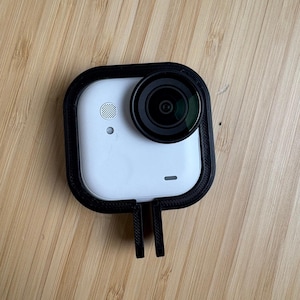 May include: A white action camera with a black lens and a black frame. The camera has a small speaker and a small light. The camera is mounted in a black frame with two prongs.