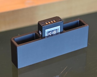 Peut inclure: Un porte-piles en plastique noir avec une seule pile à l'intérieur. La pile est noire avec des contacts dorés et le texte "LEICA" imprimé dessus.