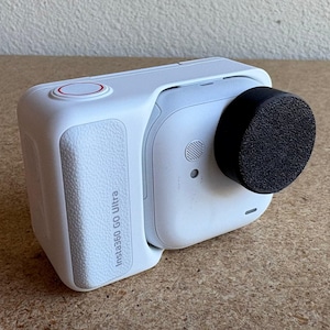 以下が含まれることがあります： 黒いレンズカバーが付いた白いInsta360 Go Ultraカメラ。カメラには赤いボタンがあり、「Insta360 Go Ultra」という文字が側面に表示されています。カメラは明るい茶色の表面にあります。