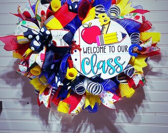 May include: A colorful back-to-school wreath with red, yellow, blue, and black ribbon, mesh, and patterned fabric. The wreath features a round sign with a red apple, pencil, and the text "Welcome to our class".