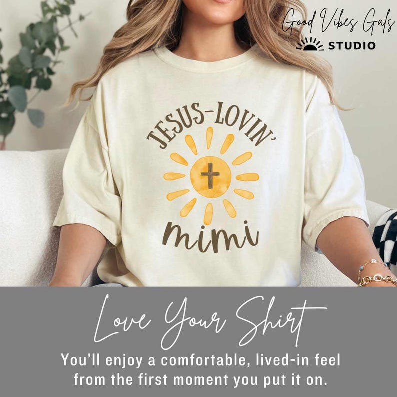 May include: Cream-colored t-shirt with the text "JESUS-LOVIN' mimi" in brown script. The design includes a yellow sun with a cross in the center. The shirt has a comfortable, lived-in feel. The brand name "Good Vibes Gals Studio" is in the top right corner.